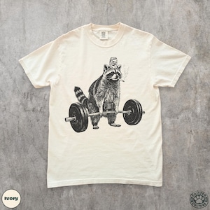 K&ouml;nnte beinhalten: Elfenbeinfarbenes T-Shirt mit einer Schwarz-Wei&szlig;-Illustration eines Waschb&auml;ren, der Gewichte hebt. Der Waschb&auml;r tr&auml;gt einen Flaschenverschluss-Hut und raucht eine Zigarette. Das T-Shirt hat einen Rundhalsausschnitt und kurze &Auml;rmel. Das Markenlogo ist sichtbar.
