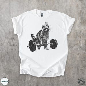 K&ouml;nnte beinhalten: Wei&szlig;es T-Shirt mit einer Schwarz-Wei&szlig;-Illustration eines Waschb&auml;ren, der eine Langhantel hebt. Der Waschb&auml;r tr&auml;gt einen Hut und raucht eine Zigarette. Das Shirt hat die Worte "Critter Culture" unten rechts.