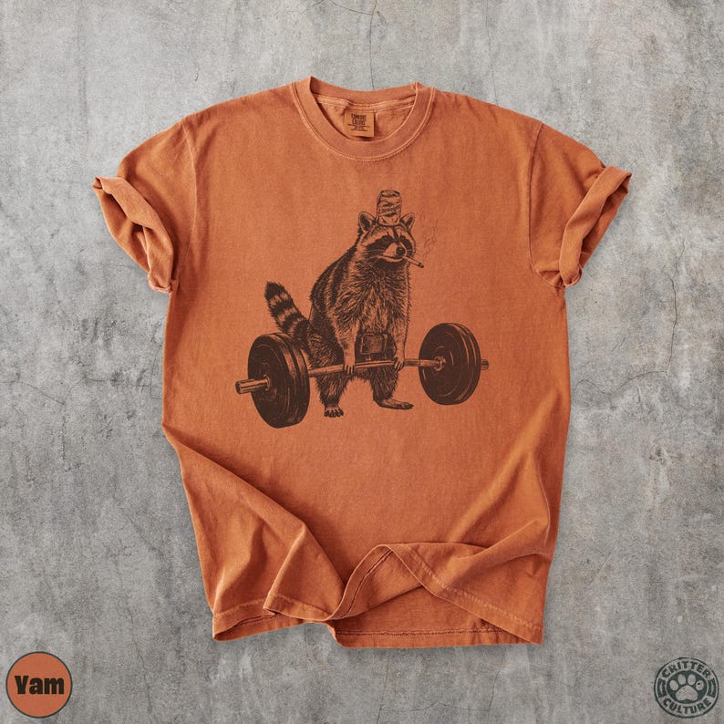 K&ouml;nnte beinhalten: Orangefarbenes T-Shirt mit einer braunen Illustration eines Waschb&auml;ren, der Gewichte stemmt. Der Waschb&auml;r tr&auml;gt einen Hut und h&auml;lt eine Langhantel. Das T-Shirt hat hochgekrempelte &Auml;rmel. Der Text "Yam" ist sichtbar.
