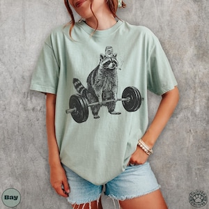 K&ouml;nnte beinhalten: Hellgr&uuml;nes T-Shirt mit einer schwarz-wei&szlig;en Illustration eines Waschb&auml;ren, der Gewichte hebt. Der Waschb&auml;r tr&auml;gt einen Flaschenverschluss-Hut und raucht eine Zigarette. Das T-Shirt hat eine lockere Passform.