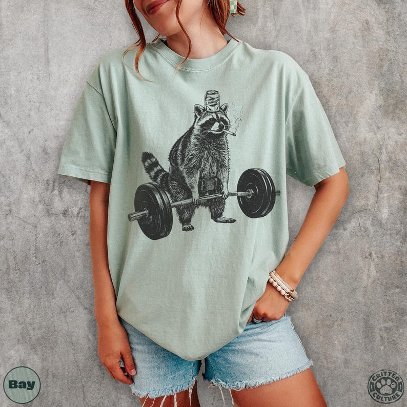 K&ouml;nnte beinhalten: Hellgr&uuml;nes T-Shirt mit einer schwarz-wei&szlig;en Illustration eines Waschb&auml;ren, der Gewichte hebt. Der Waschb&auml;r tr&auml;gt einen Flaschenverschluss-Hut und raucht eine Zigarette. Das T-Shirt hat eine lockere Passform.