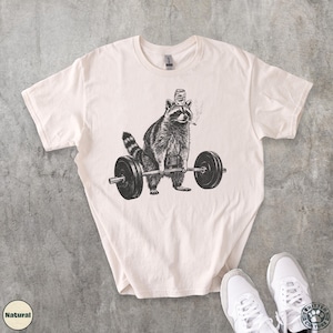 K&ouml;nnte beinhalten: Cremefarbenes T-Shirt mit einer Schwarz-Wei&szlig;-Illustration eines Waschb&auml;ren, der eine Langhantel hebt. Der Waschb&auml;r tr&auml;gt einen Hut und raucht eine Zigarette. Das Shirt hat einen Rundhalsausschnitt und kurze &Auml;rmel. Wei&szlig;e Turnschuhe sind unten rechts zu sehen.