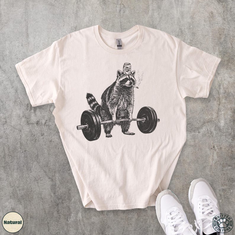 K&ouml;nnte beinhalten: Cremefarbenes T-Shirt mit einer Schwarz-Wei&szlig;-Illustration eines Waschb&auml;ren, der eine Langhantel hebt. Der Waschb&auml;r tr&auml;gt einen Hut und raucht eine Zigarette. Das Shirt hat einen Rundhalsausschnitt und kurze &Auml;rmel. Wei&szlig;e Turnschuhe sind unten rechts zu sehen.