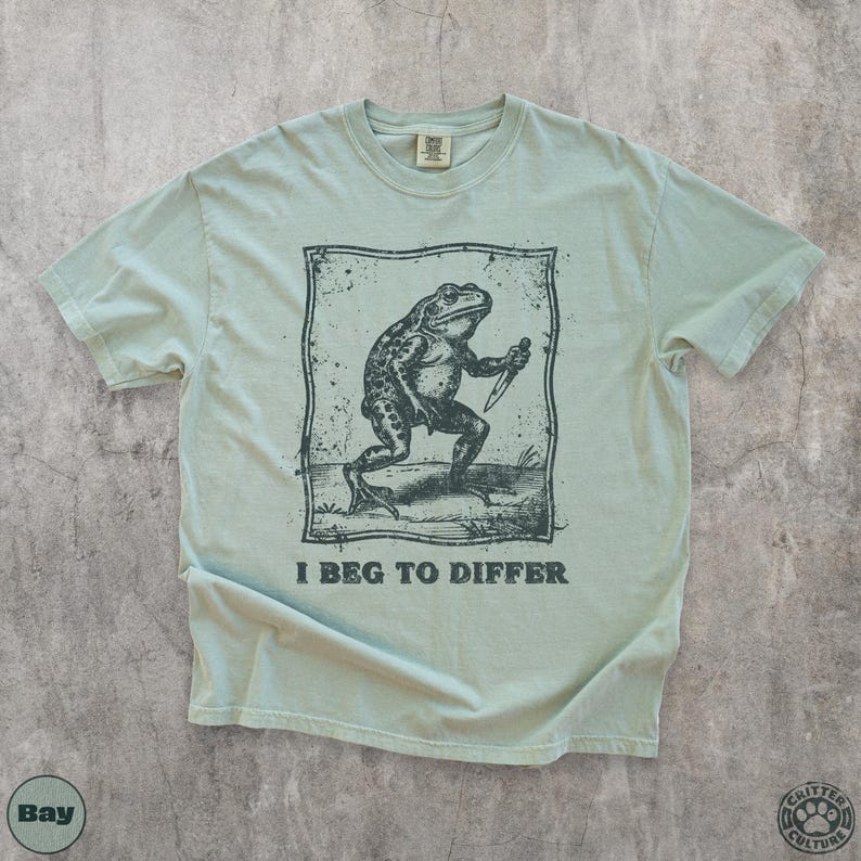 Peut inclure: T-shirt vert clair avec une illustration en noir et blanc d'une grenouille tenant un couteau, avec le texte "I Beg to Differ" en dessous. Le t-shirt a un col rond et des manches courtes. Le logo de la marque est en bas &agrave; droite.