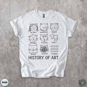 以下が含まれることがあります： 有名な芸術家のスタイルで描かれた猫の顔の黒いグラフィックが特徴の、明るい灰色のTシャツ。ダ・ヴィンチ、レンブラント、ウォーホルなどが含まれます。「HISTORY OF ART」の文字が猫のイラストの下に印刷されています。柔らかい素材のシャツです。
