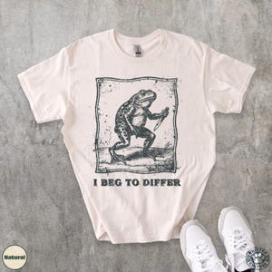 Peut inclure: T-shirt de couleur cr&egrave;me avec une illustration en noir et blanc d'une grenouille tenant un couteau, encadr&eacute;e dans un rectangle. Le texte "I BEG TO DIFFER" est imprim&eacute; en dessous. Le t-shirt est pr&eacute;sent&eacute; avec des baskets blanches.