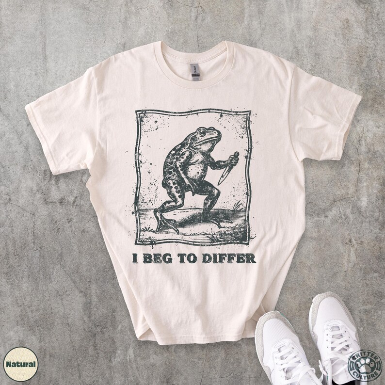 Peut inclure: T-shirt de couleur cr&egrave;me avec une illustration en noir et blanc d'une grenouille tenant un couteau, encadr&eacute;e dans un rectangle. Le texte "I BEG TO DIFFER" est imprim&eacute; en dessous. Le t-shirt est pr&eacute;sent&eacute; avec des baskets blanches.