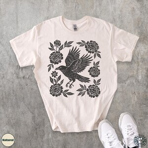 May include: Cream-colored t-shirt featuring a black crow design with outstretched wings, surrounded by floral elements. The graphic is centered on the shirt. The shirt is paired with white sneakers.