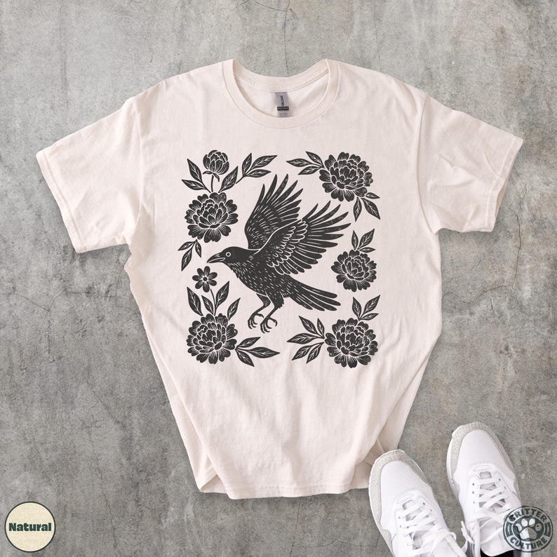 May include: Cream-colored t-shirt featuring a black crow design with outstretched wings, surrounded by floral elements. The graphic is centered on the shirt. The shirt is paired with white sneakers.