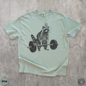 K&ouml;nnte beinhalten: Hellgr&uuml;nes T-Shirt mit einer Schwarz-Wei&szlig;-Illustration eines Waschb&auml;ren, der Gewichte hebt. Der Waschb&auml;r tr&auml;gt einen Hut und raucht eine Zigarette. Das T-Shirt hat einen Rundhalsausschnitt und kurze &Auml;rmel.