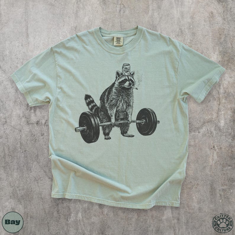 K&ouml;nnte beinhalten: Hellgr&uuml;nes T-Shirt mit einer Schwarz-Wei&szlig;-Illustration eines Waschb&auml;ren, der Gewichte hebt. Der Waschb&auml;r tr&auml;gt einen Hut und raucht eine Zigarette. Das T-Shirt hat einen Rundhalsausschnitt und kurze &Auml;rmel.