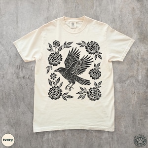 May include: Ivory-colored t-shirt featuring a black raven with outstretched wings, surrounded by floral elements. The design is centered on the shirt, with the text "Critter Culture" and "Ivory" visible. A casual, comfortable tee.