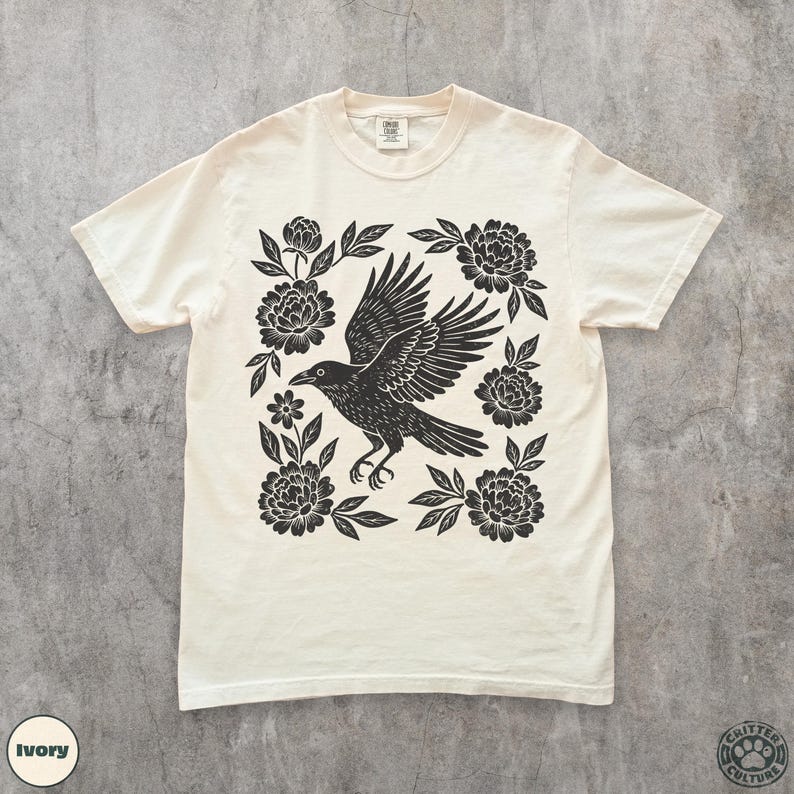 May include: Ivory-colored t-shirt featuring a black raven with outstretched wings, surrounded by floral elements. The design is centered on the shirt, with the text "Critter Culture" and "Ivory" visible. A casual, comfortable tee.