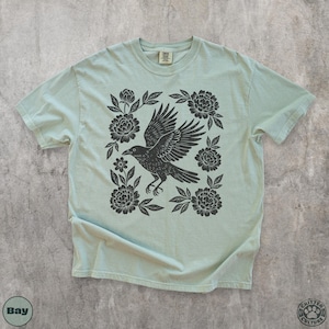 May include: A light green t-shirt featuring a black raven with outstretched wings, surrounded by floral elements. The design is centered on the shirt, with the text "Bay" and "Critter Culture" visible.