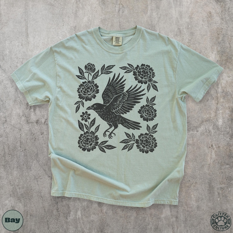 May include: A light green t-shirt featuring a black raven with outstretched wings, surrounded by floral elements. The design is centered on the shirt, with the text "Bay" and "Critter Culture" visible.