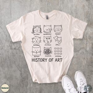 以下が含まれることがあります： クリーム色のTシャツ。ダ・ヴィンチ、ゴッホ、ウォーホルなど、有名なアーティスト風の猫の顔の黒いグラフィックが特徴です。「HISTORY OF ART」の文字が猫のイラストの下にプリントされています。白いスニーカーが右下に写っています。