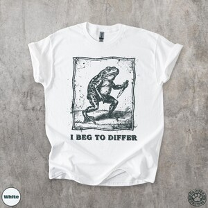 Peut inclure: T-shirt blanc avec une illustration en noir et blanc d'une grenouille tenant un couteau, avec le texte "I Beg to Differ". Le t-shirt a un col rond et des manches courtes.