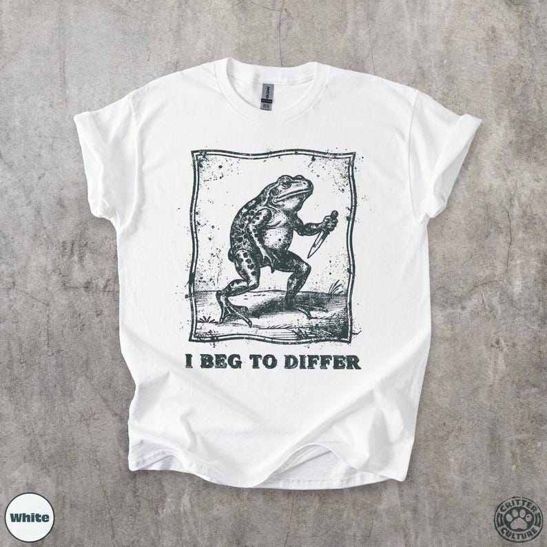 Peut inclure: T-shirt blanc avec une illustration en noir et blanc d'une grenouille tenant un couteau, avec le texte "I Beg to Differ". Le t-shirt a un col rond et des manches courtes.