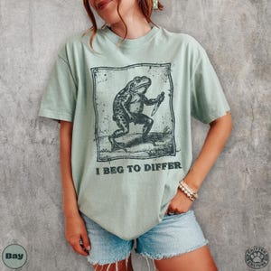 Peut inclure: T-shirt vert sauge avec une illustration en noir et blanc d'une grenouille tenant un couteau, et le texte "I BEG TO DIFFER". Le t-shirt a une coupe ample et des manches courtes.