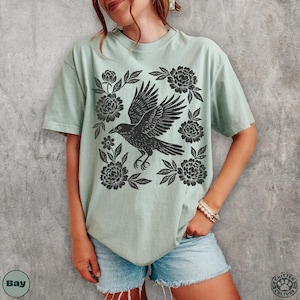 May include: A light green t-shirt featuring a black crow design surrounded by floral elements. The crow is depicted with outstretched wings, and the flowers are stylized. The shirt is a comfortable, casual style.