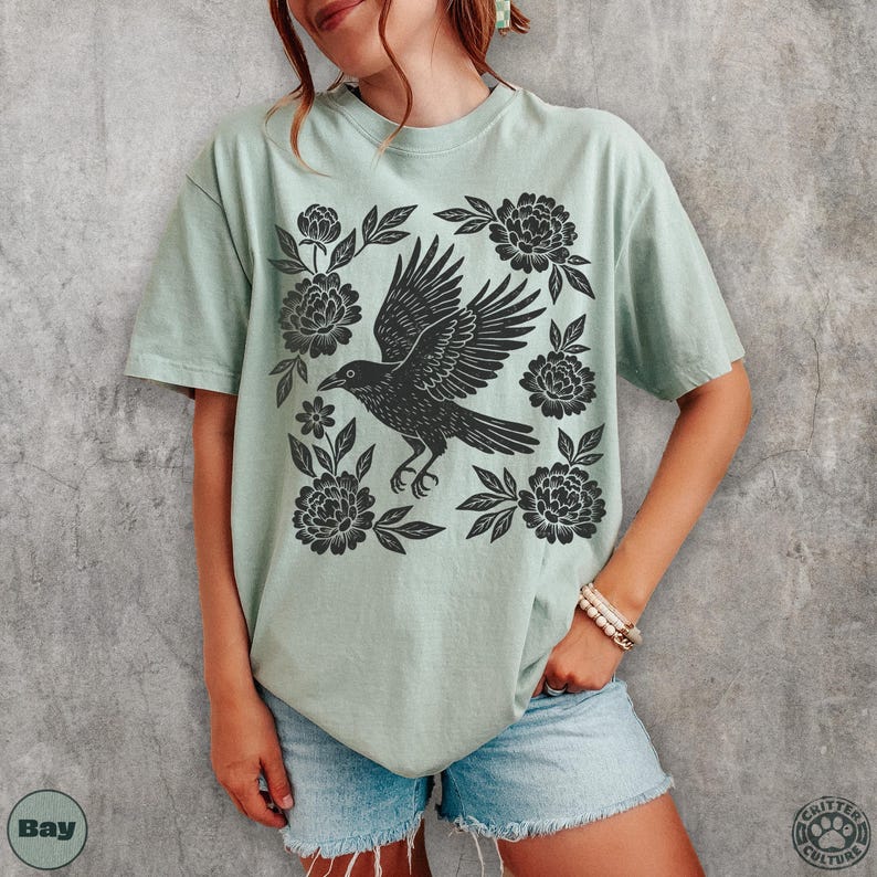 May include: A light green t-shirt featuring a black crow design surrounded by floral elements. The crow is depicted with outstretched wings, and the flowers are stylized. The shirt is a comfortable, casual style.