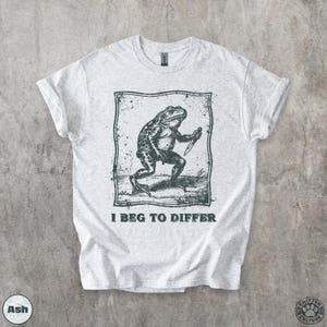 Peut inclure: T-shirt gris clair avec une illustration en noir et blanc d'une grenouille tenant un couteau, dans un cadre rectangulaire. Le texte "I BEG TO DIFFER" est en dessous. Les manches du t-shirt sont retrouss&eacute;es.