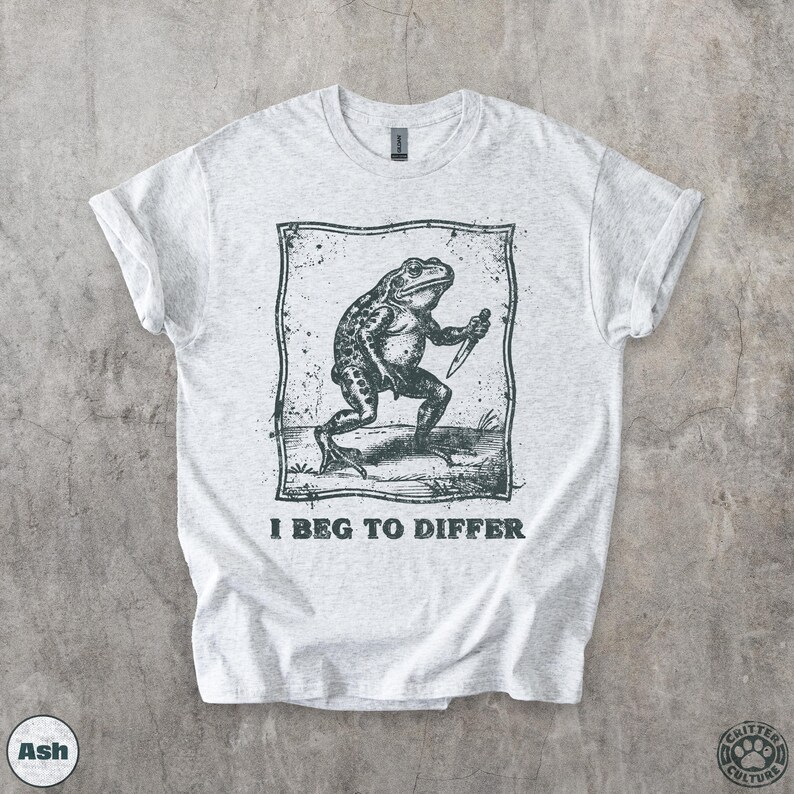 Peut inclure: T-shirt gris clair avec une illustration en noir et blanc d'une grenouille tenant un couteau, dans un cadre rectangulaire. Le texte "I BEG TO DIFFER" est en dessous. Les manches du t-shirt sont retrouss&eacute;es.
