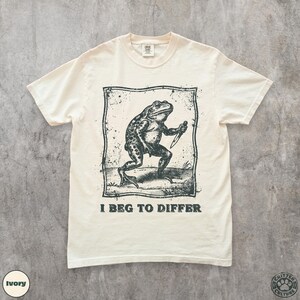 Peut inclure: T-shirt ivoire avec une illustration en noir et blanc d'une grenouille tenant un couteau, encadr&eacute;e dans un rectangle. Le texte "I BEG TO DIFFER" est imprim&eacute; en dessous. Le t-shirt a un col rond et des manches courtes.