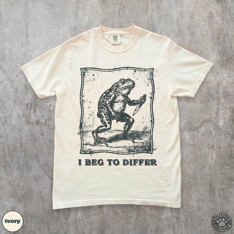 Peut inclure: T-shirt ivoire avec une illustration en noir et blanc d'une grenouille tenant un couteau, encadr&eacute;e dans un rectangle. Le texte "I BEG TO DIFFER" est imprim&eacute; en dessous. Le t-shirt a un col rond et des manches courtes.