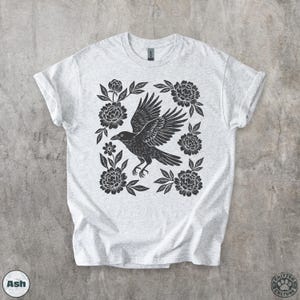May include: Light gray t-shirt featuring a black raven with outstretched wings, surrounded by black floral elements. The design is centered on the shirt. The shirt has rolled-up sleeves. The word "Ash" is printed on the bottom left.