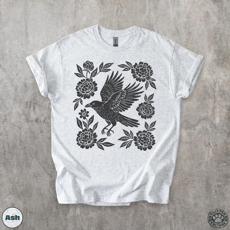 May include: Light gray t-shirt featuring a black raven with outstretched wings, surrounded by black floral elements. The design is centered on the shirt. The shirt has rolled-up sleeves. The word "Ash" is printed on the bottom left.