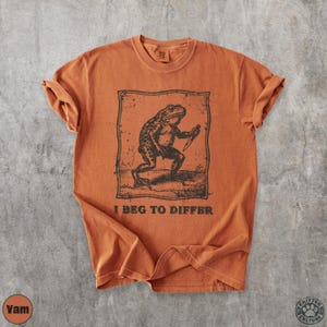 Peut inclure: T-shirt orange br&ucirc;l&eacute; avec une illustration en noir et blanc d'une grenouille tenant un couteau, avec le texte "I BEG TO DIFFER". Le t-shirt a des manches retrouss&eacute;es et un col rond.