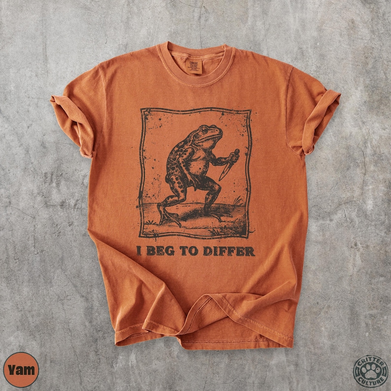 Peut inclure: T-shirt orange br&ucirc;l&eacute; avec une illustration en noir et blanc d'une grenouille tenant un couteau, avec le texte "I BEG TO DIFFER". Le t-shirt a des manches retrouss&eacute;es et un col rond.