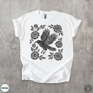 May include: White t-shirt featuring a black graphic of a raven with outstretched wings, surrounded by floral elements. The design is centered on the shirt, creating a striking contrast against the white fabric. The shirt has a crew neck and short sleeves.