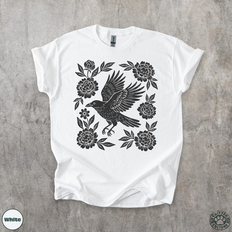 May include: White t-shirt featuring a black graphic of a raven with outstretched wings, surrounded by floral elements. The design is centered on the shirt, creating a striking contrast against the white fabric. The shirt has a crew neck and short sleeves.