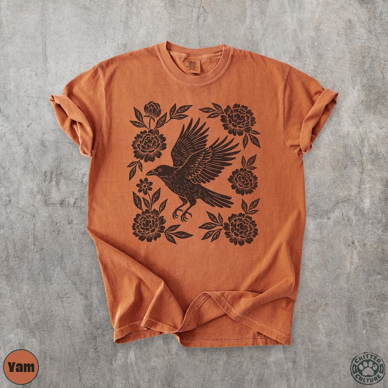 May include: Burnt orange t-shirt featuring a black raven with outstretched wings, surrounded by floral elements. The design is centered on the shirt, with rolled sleeves. The shirt has a vintage, slightly faded appearance.
