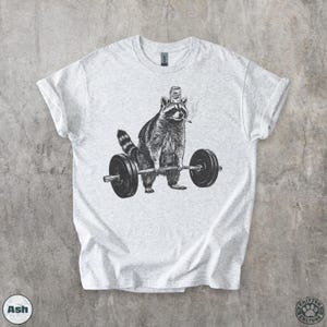K&ouml;nnte beinhalten: Hellgraues T-Shirt mit einer Schwarz-Wei&szlig;-Illustration eines Waschb&auml;ren, der Gewichte hebt. Der Waschb&auml;r tr&auml;gt eine Dose auf dem Kopf und raucht eine Zigarette. Das Shirt hat den Text "Critter Culture" unten rechts.