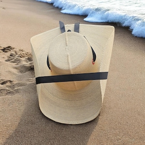 May include: A large, beige straw hat with a smaller, round hat inside, secured by a black band. The hats are on a sandy beach near the sea. The hat has a black band and red accents.
