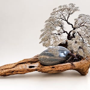 May include: A wire sculpture of a tree with silver-colored metallic branches and a dark brown trunk, anchored to a smooth, oval-shaped gray stone. The entire assembly rests upon a piece of textured, weathered natural driftwood.