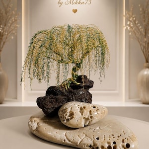 May include: A wire sculpture of a weeping willow tree with green and gold metallic strands, anchored to a dark textured rock. The sculpture rests on a large, porous, light-colored stone. Text above reads: handmade with love by Mikka 73.