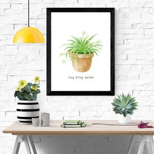 May include: A framed watercolor print of a spider plant in a terracotta pot. The print has a black frame and the text "itsy bitsy spider" below the plant. The print is on a white brick wall.
