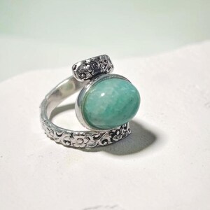 Könnte beinhalten: Ein silberner Ring mit einem großen, ovalen, hellgrünen Edelstein. Der Ring hat ein verziertes, florales Design am Band und ein kleineres, dekoratives Element über dem Edelstein. Der Ring ist vor einem weißen Hintergrund platziert.