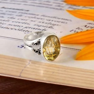 Könnte beinhalten: Ein silberner Ring mit einem ovalen, durchscheinenden, gelb-grünen Edelstein. Der Edelstein hat goldfarbene Einschlüsse. Der Ring liegt auf einem Buch mit floralem Akzent.
