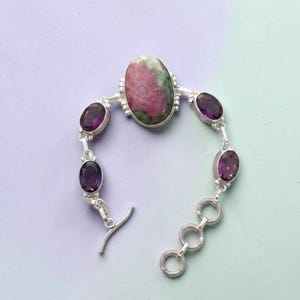 Könnte beinhalten: Ein silbernes Armband mit einem ovalen, rosa-grünen Mittelstein und vier ovalen, tiefvioletten Edelsteinen. Das Armband hat einen Knebelverschluss. Der Hintergrund ist helllila.