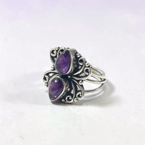 Könnte beinhalten: Ein Silberring mit zwei vertikal angeordneten, tropfenförmigen violetten Amethyst-Edelsteinen. Das Metallband zeigt komplizierte Schnörkel und Filigranverzierungen um die Steine herum, mit einem mehrsträngigen Ringschienen-Design an den Seiten.