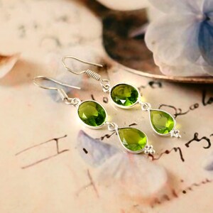 Könnte beinhalten: Ein Paar silberne Ohrringe mit drei grünen Peridot-Edelsteinen. Die Ohrringe haben ein Haken-Design und verfügen über ovale und tropfenförmige Steine. Die Edelsteine sind von leuchtend grüner Farbe und in Silbermetall gefasst.