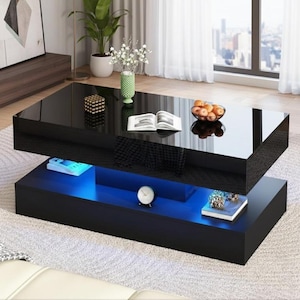 May include: A modern black coffee table with a glossy finish. The table has two levels, with blue LED lighting illuminating the space between them. Decorative items like a clock, books, and a bowl of fruit are on display.