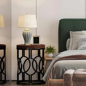 May include: A bedside table with a dark brown top and black metal base, supporting a lamp with a cream shade and a decorative ceramic base. A small framed picture and a potted plant are also on the table. The bed has a green headboard.