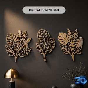 Op de afbeelding: Drie goudkleurige wanddecoraties met botanische bladmotieven, waaronder monstera- en varenvormen. De tekst "DIGITAL DOWNLOAD" staat bovenaan. Deze decoratieve items zijn op een rij geplaatst tegen een donkergrijze muur.