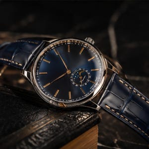 May include: A sophisticated wristwatch featuring a dark blue dial with gold hands and markers. The watch has a silver case and a blue leather strap with a crocodile skin effect. The watch is on a dark leather surface.